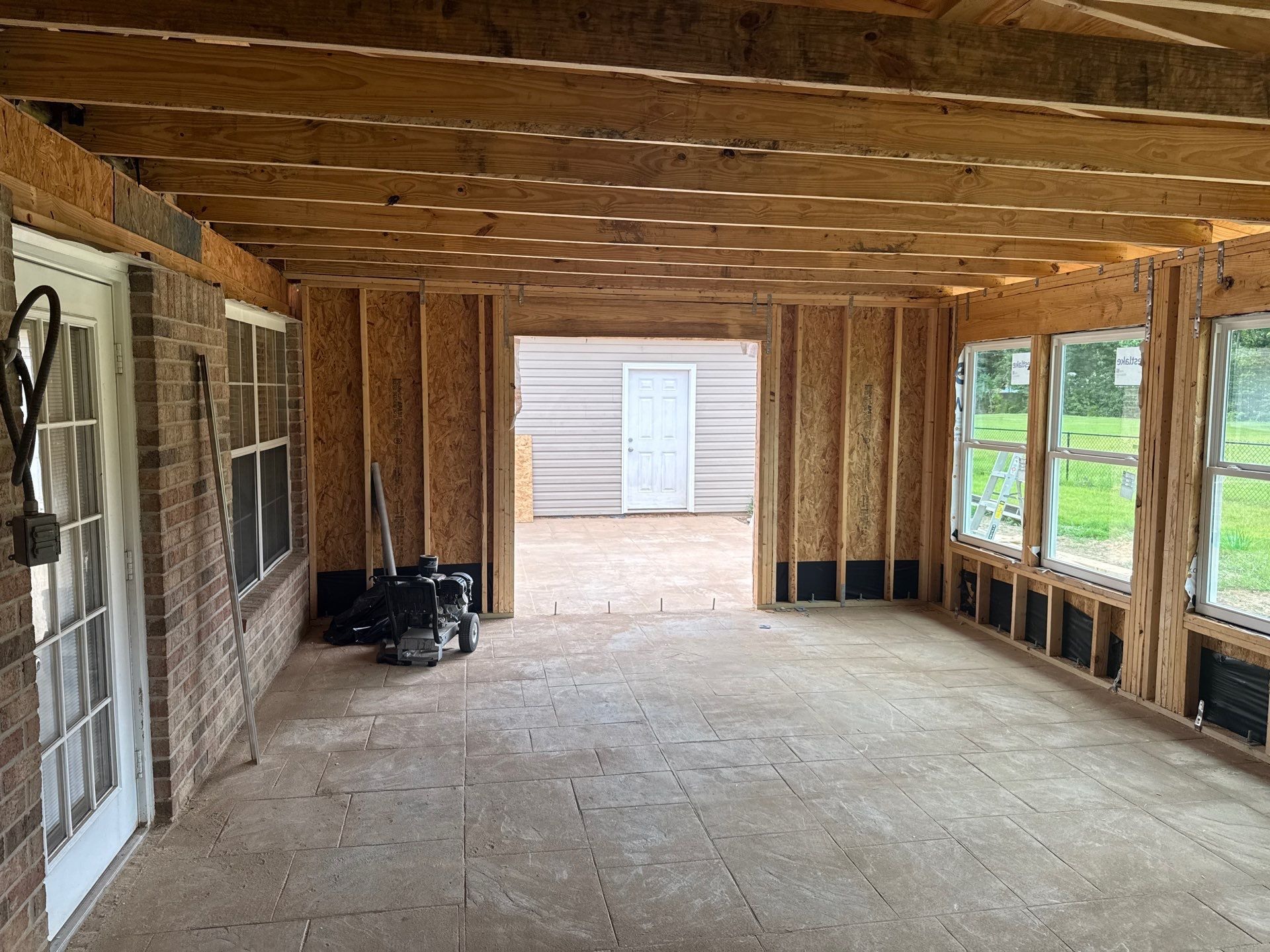 In-progress room addition interior showing new stud walls, exposed ceiling joists, sheathing, sill flashing, and newly set windows over a tiled slab